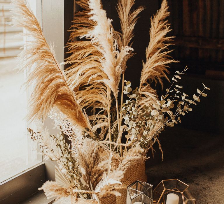 Large pampas grass stems and dried leaves in woven baskets on concrete floor next to metal and glass candle holders with pillar candles for wedding at Anran Devon