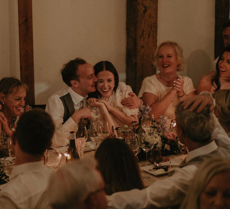 Bride in lace puffed sleeve Daalarna wedding dress is hugged by groom in white shirt and green waistcoat as they sit as wedding breakfast table surrounded by wedding guests at Loseley Park