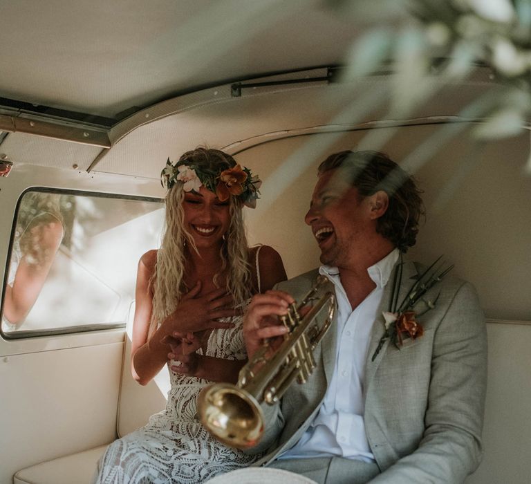 Bride in white lace boho wedding dress and tropical flowers crown smiles as groom in white shirt and linen suit laughs holding trumpet whilst they sit in the back of campervan for birds of paradise wedding inspiration