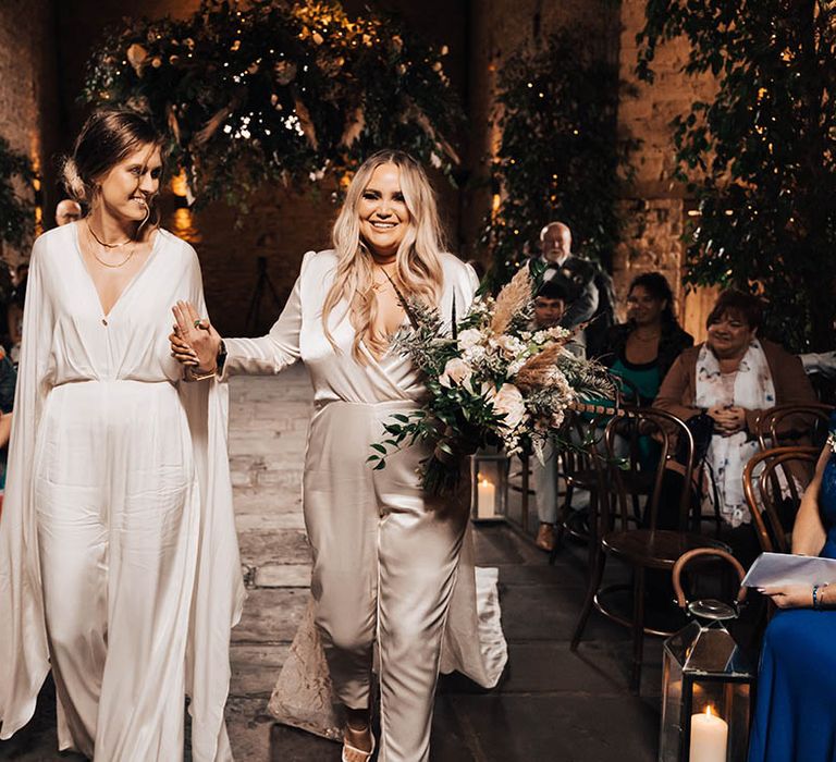 Brides walk down the aisle with one another as blonde bride holds floral bouquet and wedding guests look on whilst smiling