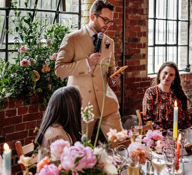 Groom in double breasted linen suit and burgundy tie holds microphone and phone during wedding breakfast in exposed brick room with florals and candles at Loft Studios London