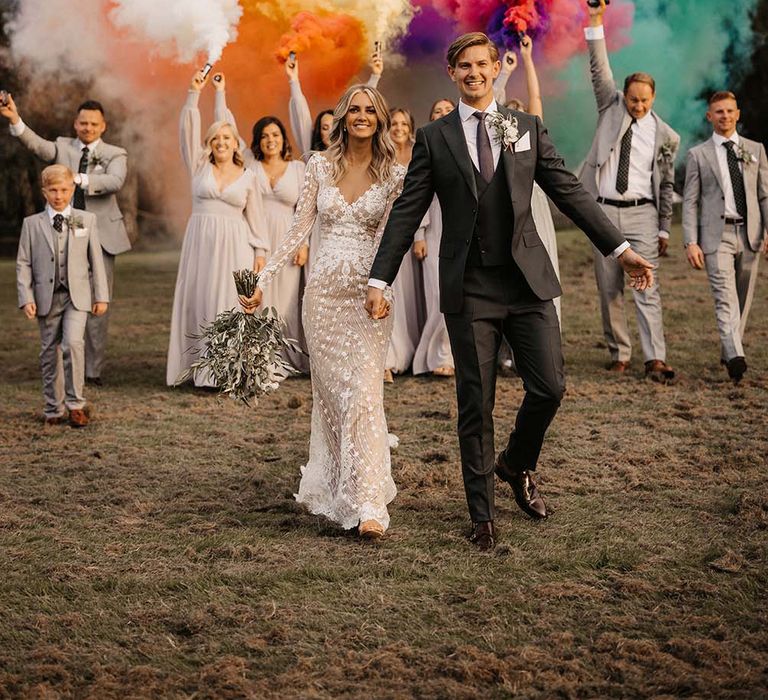 Bride in a fitted lace wedding dress holding hands with her groom in a dark grey suit as they are followed by their wedding party holding a rainbow or coloured smoke bombs