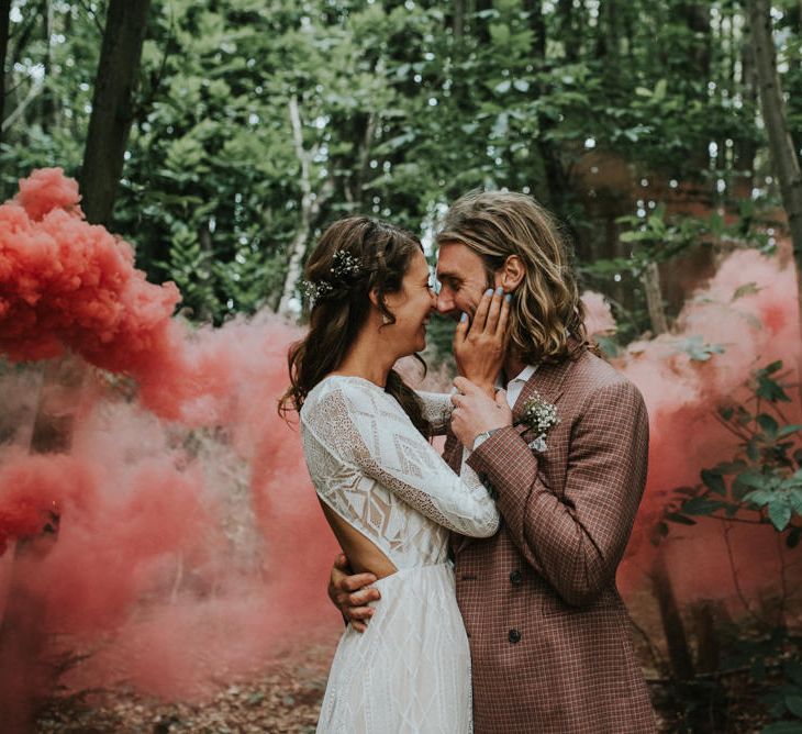 Boho bride in a lace wedding dress with long sleeve embracing her husbands face in a brown check suit with a red smoke bomb going off in the background