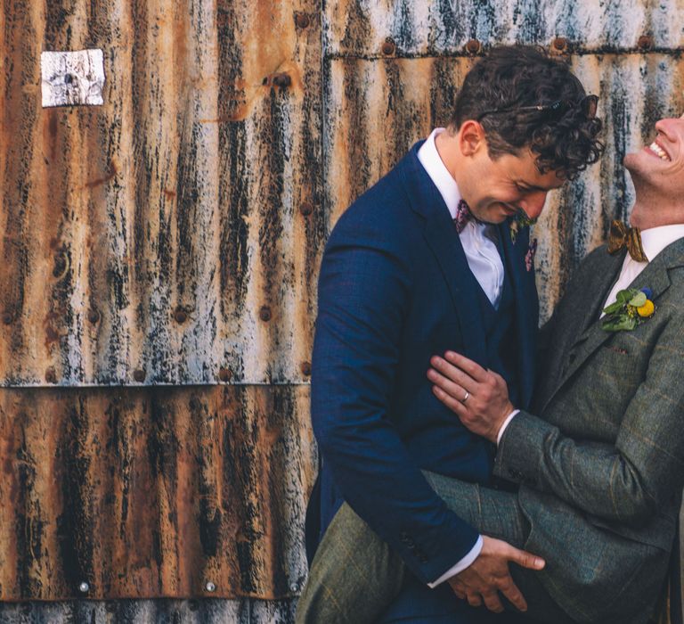 Grooms laugh with one another on their wedding day in front of industrial style wall | Story + Colour