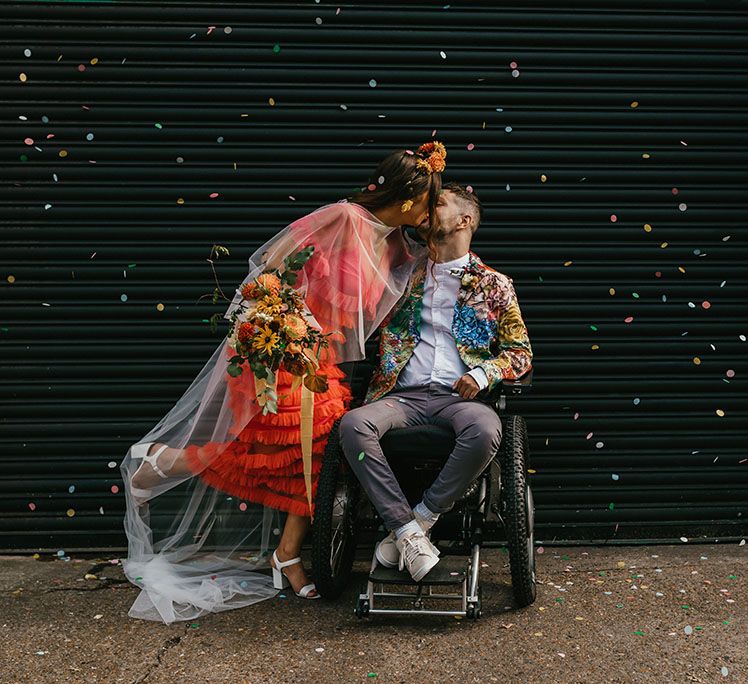 City elopement in Shoreditch with bride in coral wedding dress kissing her disabled groom in a brightly coloured blazer 