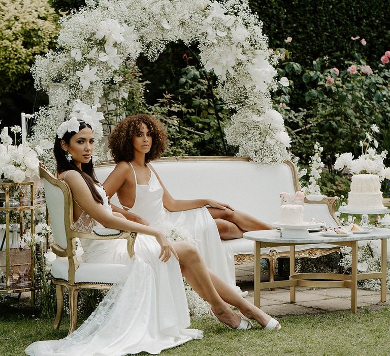 Brides in satin and lace wedding dresses sitting at their white on white outdoor tea party with gysophila flower arch and iced cakes 