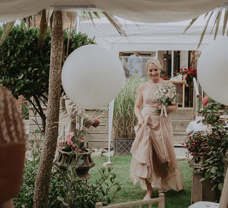 Bridesmaid in a pink chiffon dress walking down the aisle towards the intimate back garden wedding ceremony 