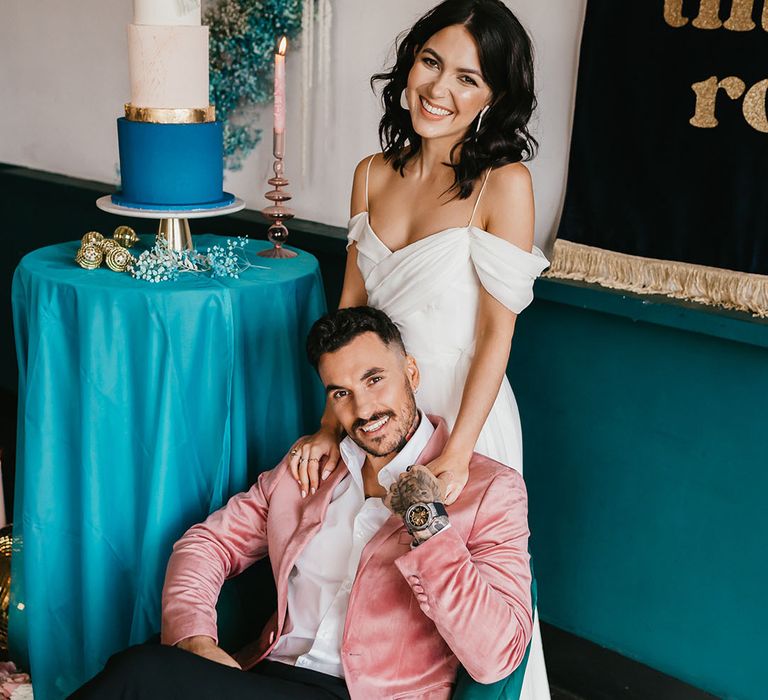 Groom in black trousers, white shirt and pink velvet jacket sitting on a teal velvet chair with his bride in a cold shoulder wedding dress standing behind him 