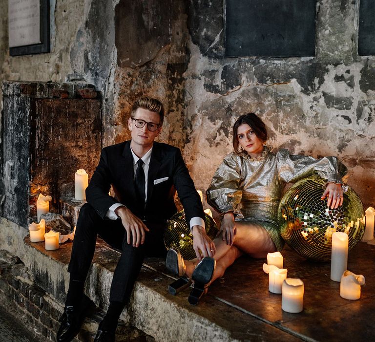 Groom in a black tie & suit with a white shirt and Bride in a croc printed gold wedding dress sitting amongst candles and disco balls