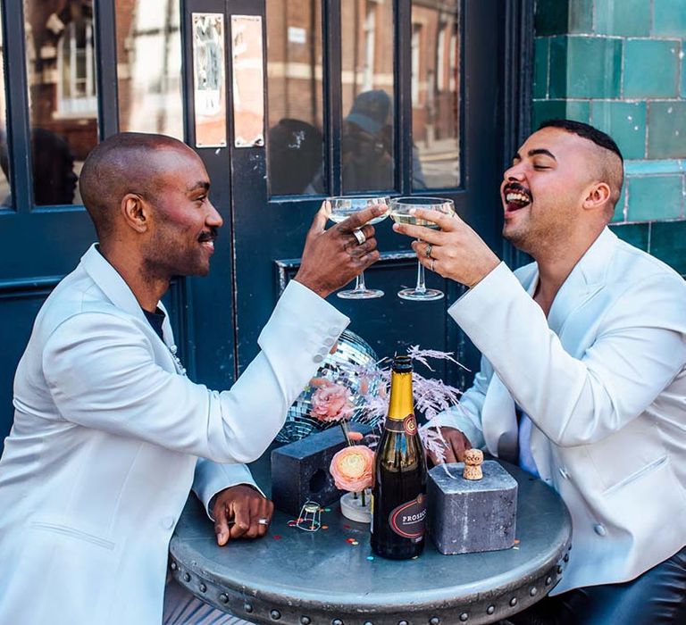 Two grooms enjoying champagne at their city elopement in white blazers with silver disco balls 