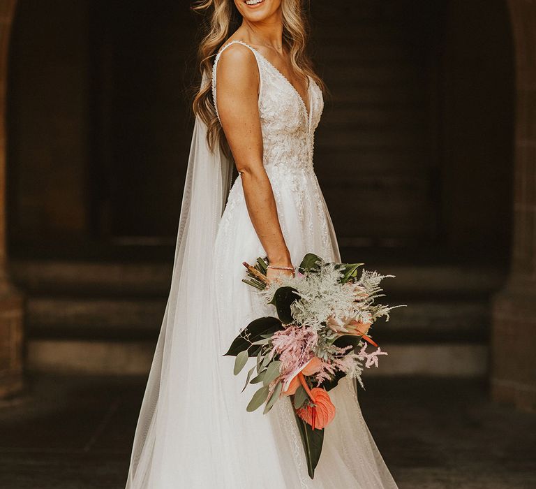 Bride wears plunging lace wedding gown and her hair in loose natural curls whilst holding tropical inspired wedding bouquet