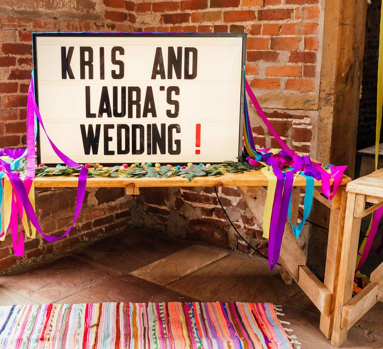 Lightbox wedding sign reading 'Kris and Laura's Wedding!' with streamers and colourful rug for barn wedding 