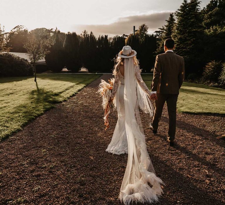 Bride in boho wedding outfit, bridal hat and fur trimmed veil holding white and orange boho bridal bouquet walks through gardens holding hands with groom in brown tweed suit