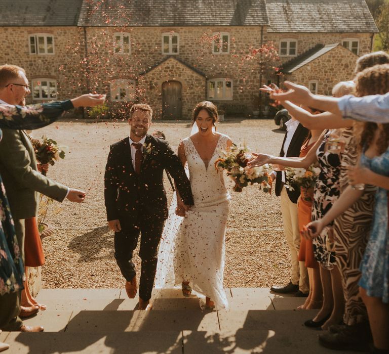 Groom in dark suit and burgundy tie holds hands with bride in lace wedding dress walking up stone steps as guests throw pink confetti