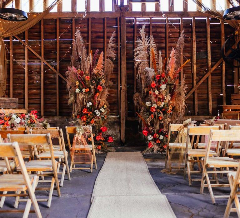 Aisle at barn wedding with rustic orange and white pampas grass floral installations with wooden chairs and fairy lights