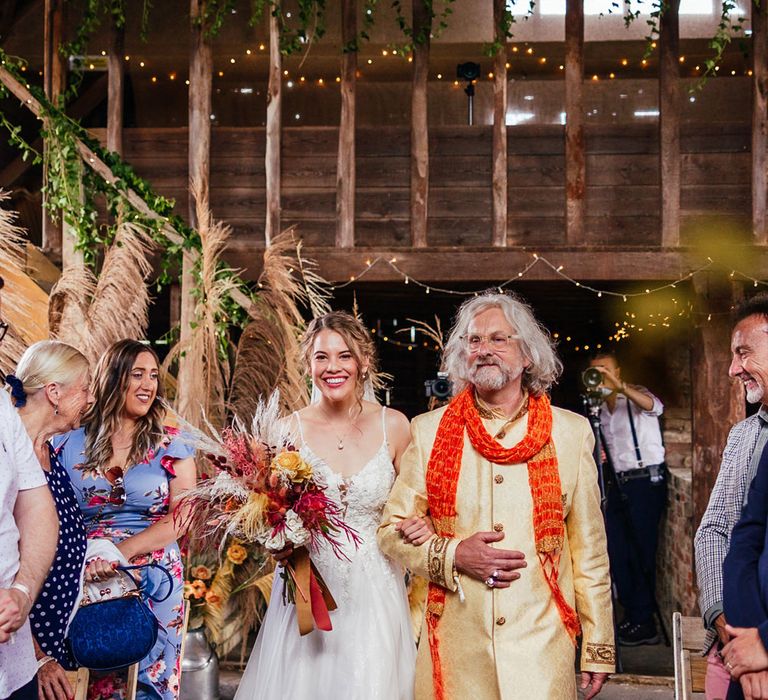 Bride in lace and tulle wedding dress holding mixed dried floral bouquet walks down the aisle arm in arm with wedding guest in yellow coat and orange scarf during barn wedding ceremony 