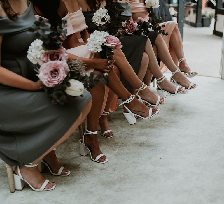 Bridesmaids wear tea length dresses whilst holding pastel bouquets and wearing white strappy block heeled sandals 