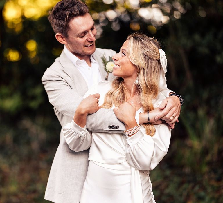 Groom in grey suit smiles whilst hugging bride with blonde curled hair, hair bow and drop pearl earrings