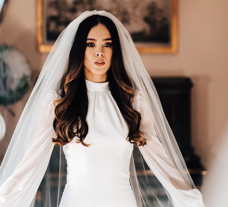 Bride with long wavy hair wearing a long sleeve fitted wedding dress and cathedral length veil 