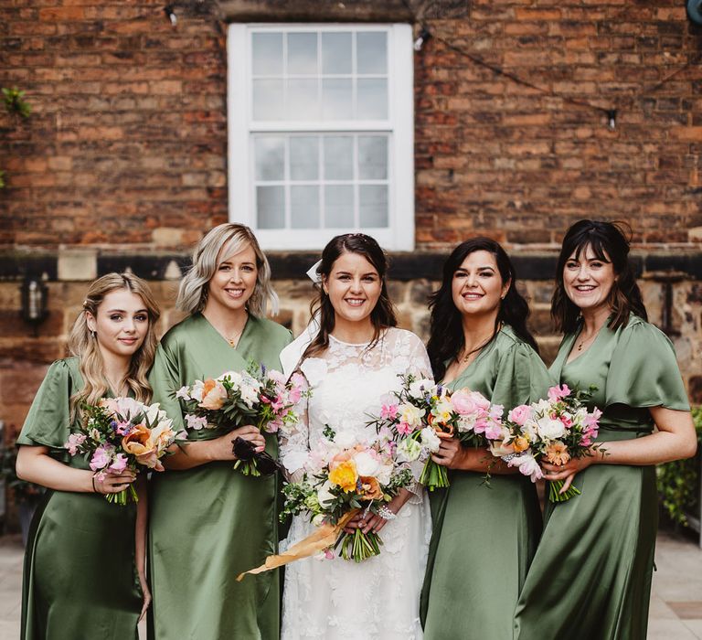 Bride in white lace wedding dress stands with bridesmaids in green satin wrap bridesmaid dresses all holding colourful bouquets