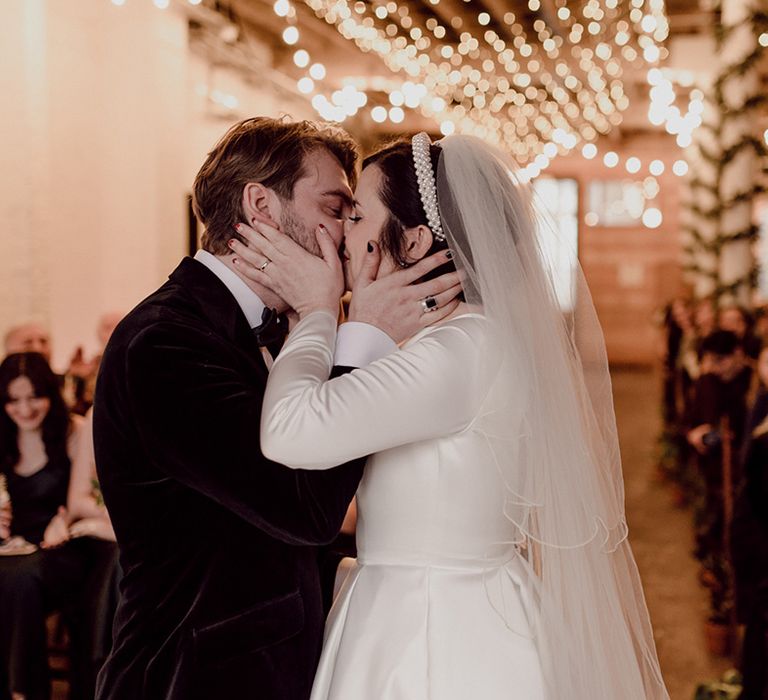 Bride and groom share their first kiss as a married couple under fairy lights and decorated venue with vines 