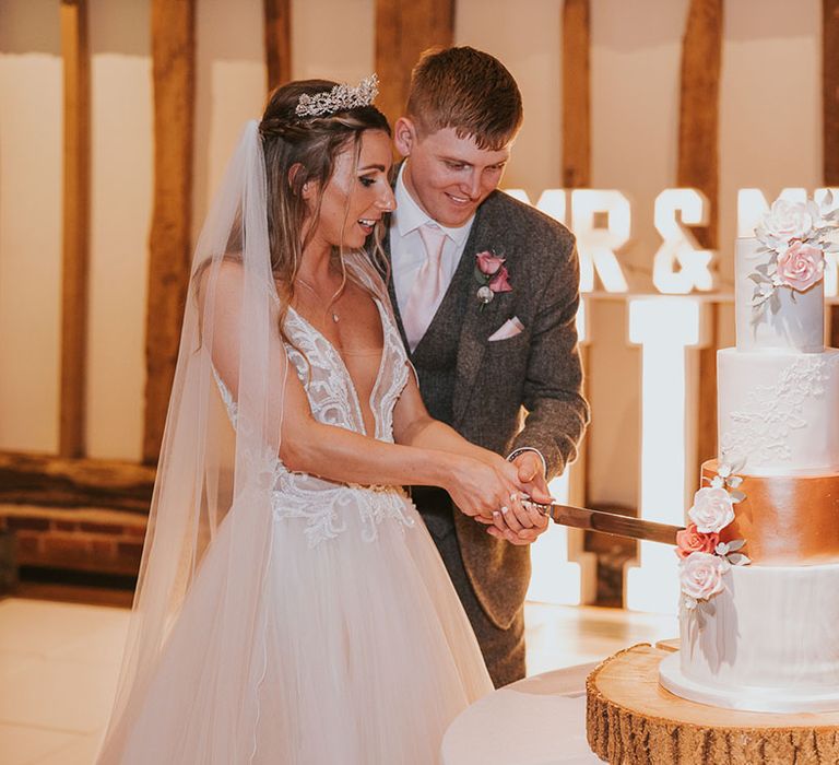 Bride and groom cut into four-tier wedding cake with roses and rose gold section in front of light-up sign 