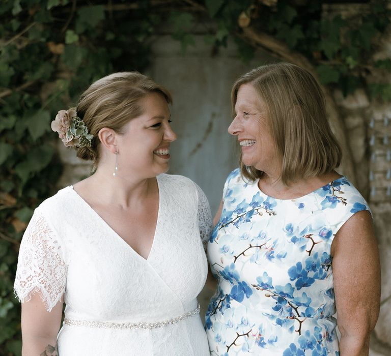 Bride in lace wedding jumpsuit and diamanté belt stands with mother in blue and white floral dress 