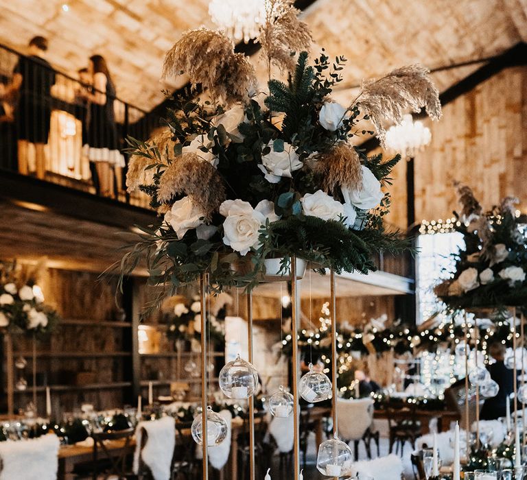 Tall white rose and dried flower centrepieces with hanging glass candles and pinecone runner with white candles table setting