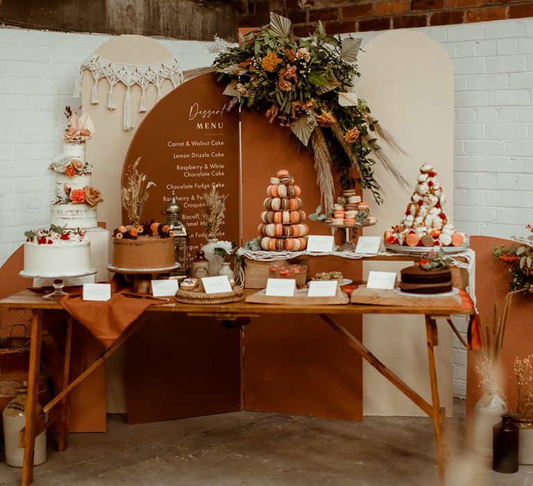 DIY dessert table with macrame, dried flowers and brown and orange sign with dessert menu featuring display of cakes, macarons and profiteroles 