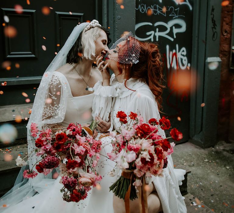 Confetti moment with two brides kissing in a short wedding dress long veil and long sleeve wedding dress and birdcage veil with pearls 