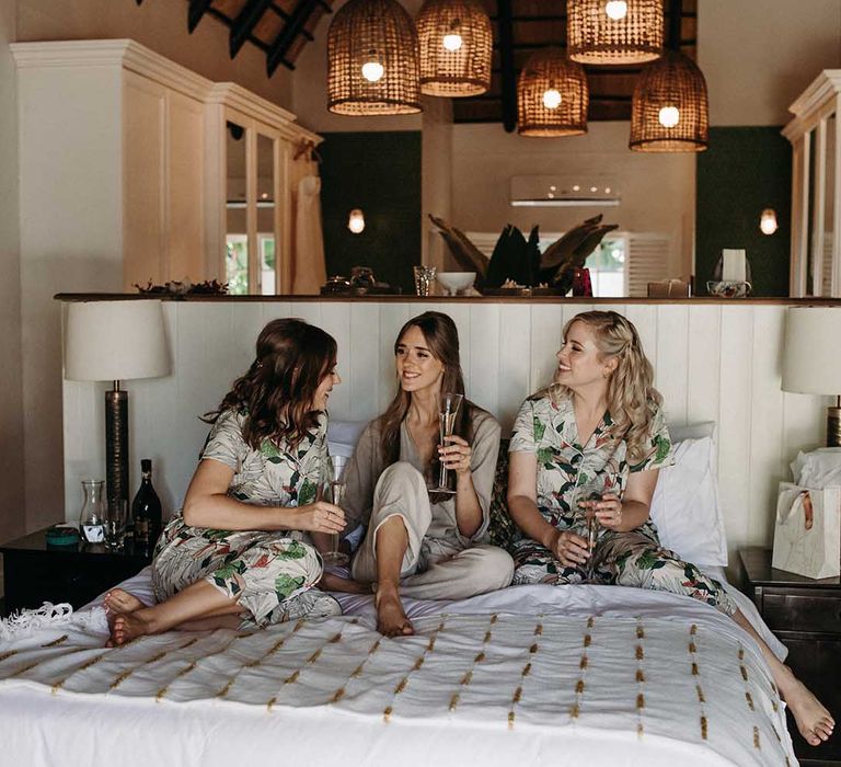 Bride in brown pyjamas and bridesmaids in leaf pattern pyjamas have a glass together on the bed before the wedding 