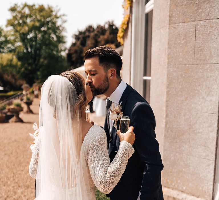 Groom in blue suit and bride in embellished dress drink champagne together outside Pennard House venue