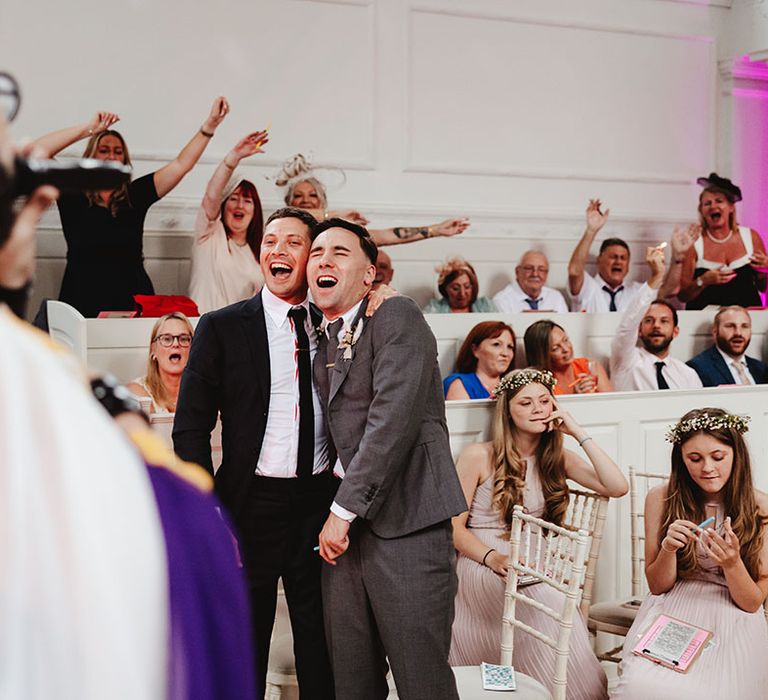 Grooms in black and grey suits have their arms around each other as they enjoy the drag entertainment 