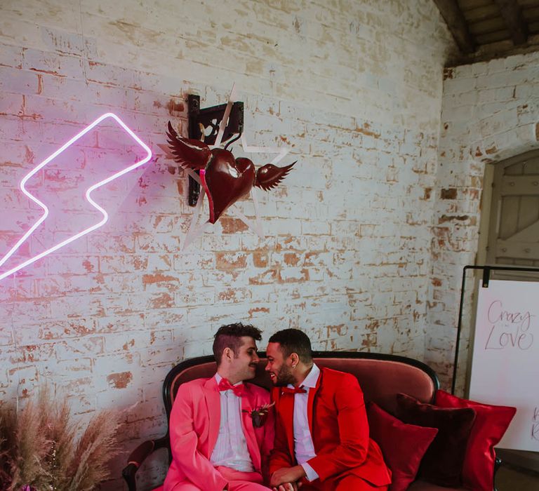 two grooms in pink and red suits sitting on an ornate velvet couch under a pink neon lightning bolt sign and heart with wings 