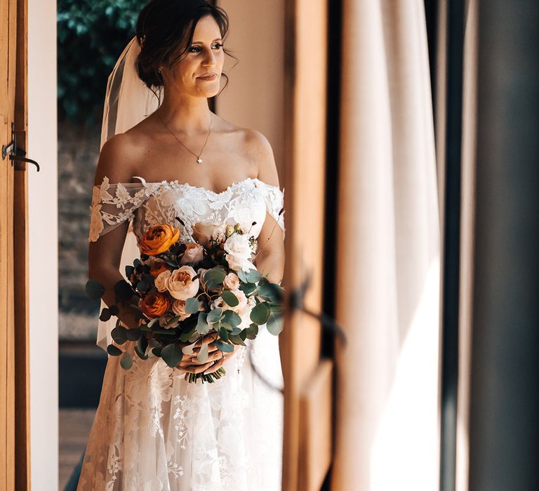 Bride with dark hair styled in an updo wearing floral lace off the shoulder wedding dress holding orange and white bouquet 