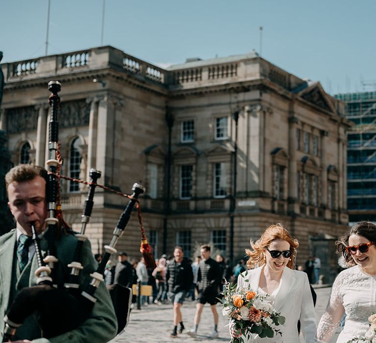 Brides hold hands and walk with one another along the Royal Mile in Edinburgh as family member plays bagpipes 