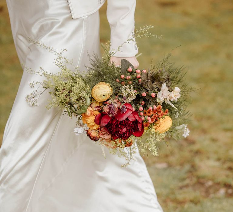 Bride in a satin slip wedding dress holding a red, orange and yellow wedding bouquet with ranunculus 