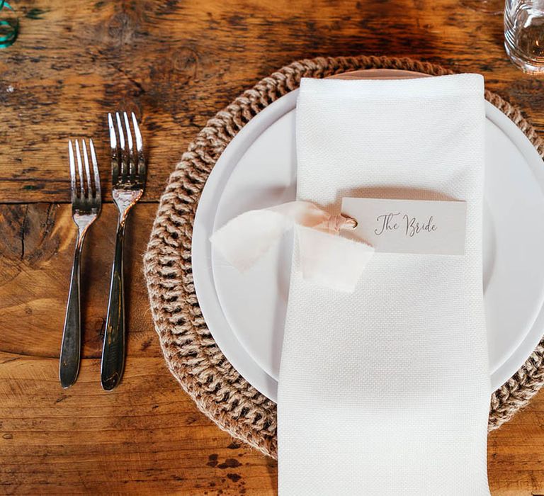 White plates on wicker place mats with white napkins and white place name cards with pink ribbon 