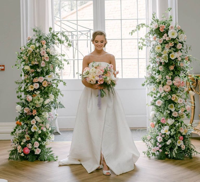 Bride stands inbetween pastel flower columns by Grace Florals holding a matching bouquet in a strapless wedding dress with a headband 