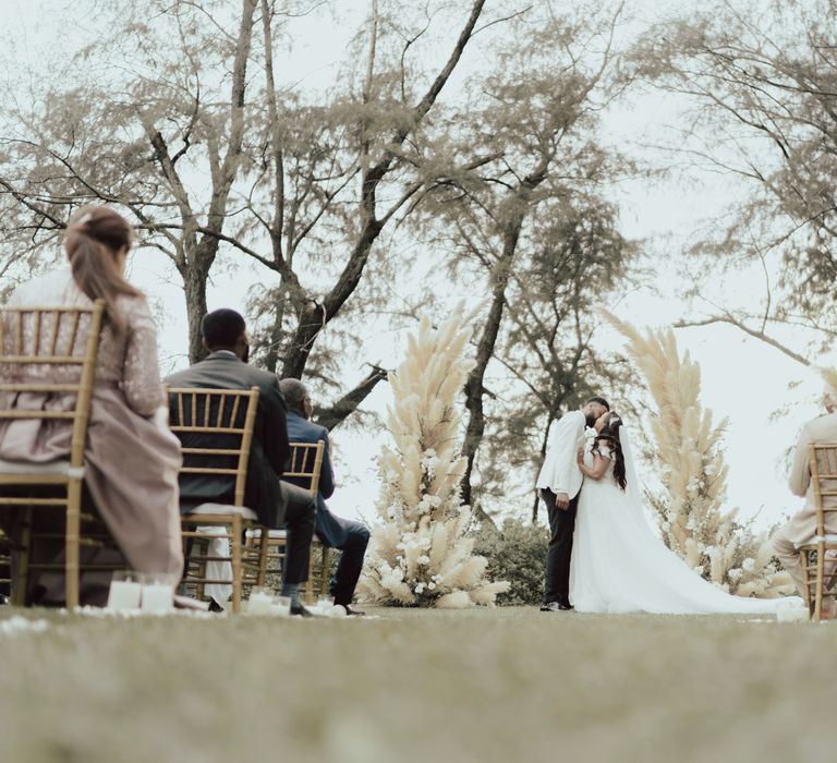 Bride & groom during their first kiss outdoors in Phuket surrounded by pampas grass arch 