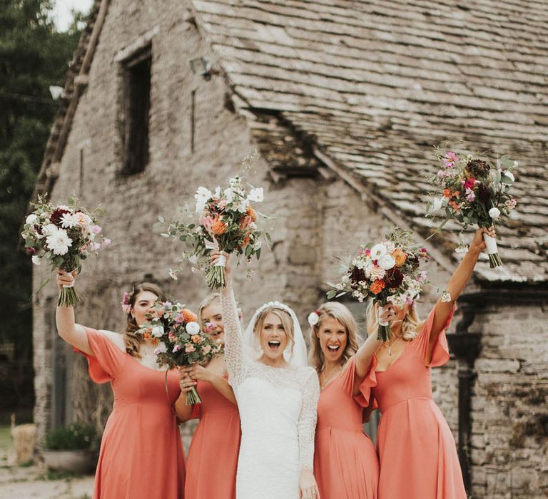 Bride in long sleeve wedding dress lifts her bouquet with the bridesmaids in coral bridesmaid dresses