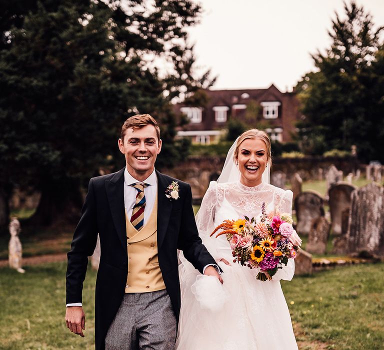 Groom in grey trousers with a yellow waistcoat and striped tie with bride in a long sleeve delicate lace wedding dress