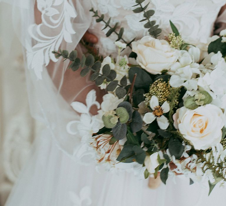 Bride in an off the shoulder sheer sleeve sweetheart lace wedding dress holding a white flower bouquet with roses 