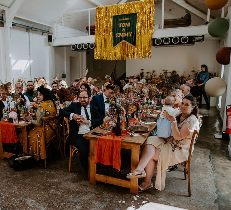 Gold streamers hang from ceiling with velvet banner as wedding guests sit at wooden banquet tables 