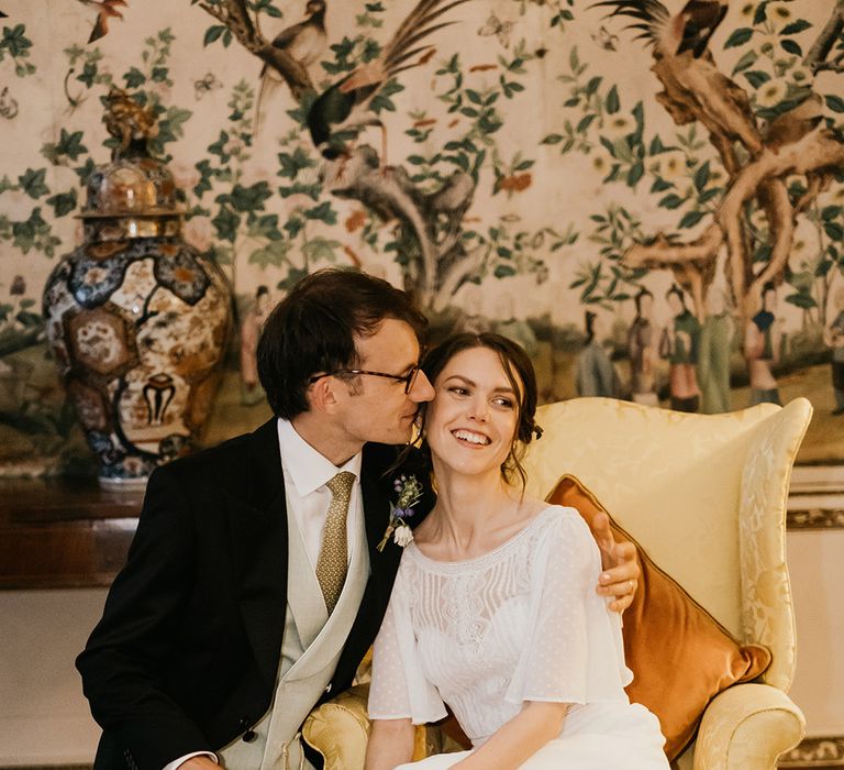 Bride in batwing sleeve lace detail wedding dress and groom in three piece suit with sage green waistcoat and tie embracing on yellow armchair with burnt orange pillow