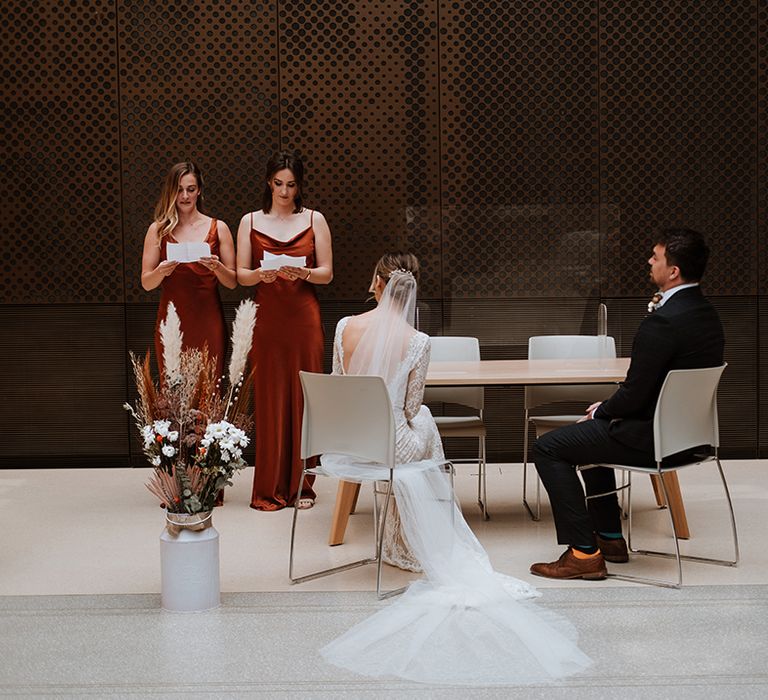 Bridesmaids in burnt orange bridesmaid dresses stand in front of bride & groom with floral bouquets to the floor