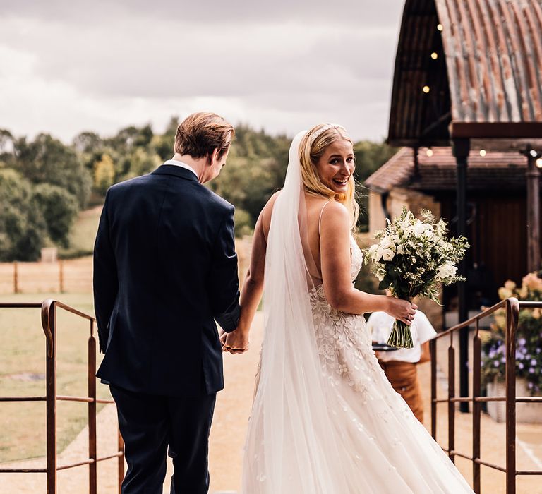 Groom in a dark blue suit walks holding hands with the bride in an appliqué wedding dress 