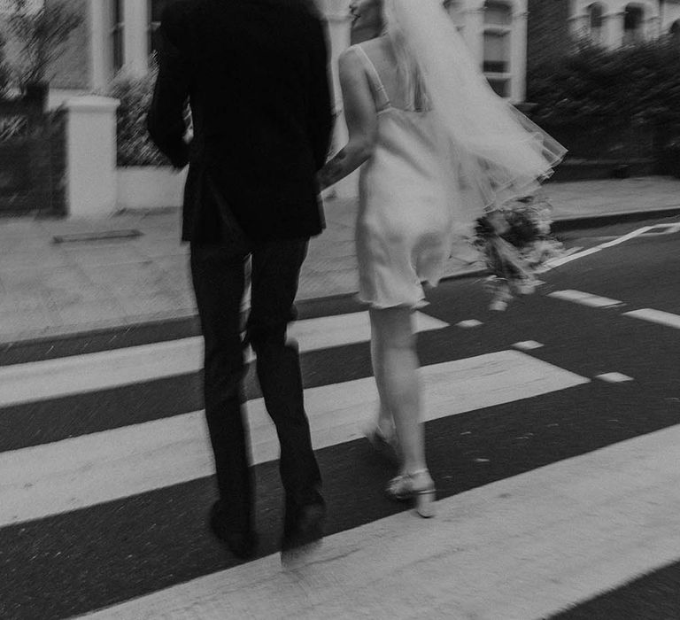 Bride in short satin dress with feather trimmed neckline and groom in dark tux with bowtie holding hands and walking