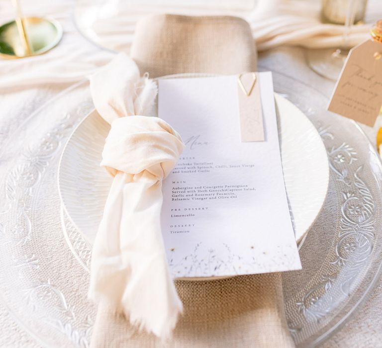 Patterned clear charger plates with white plates and a blush napkin with the wedding menu 