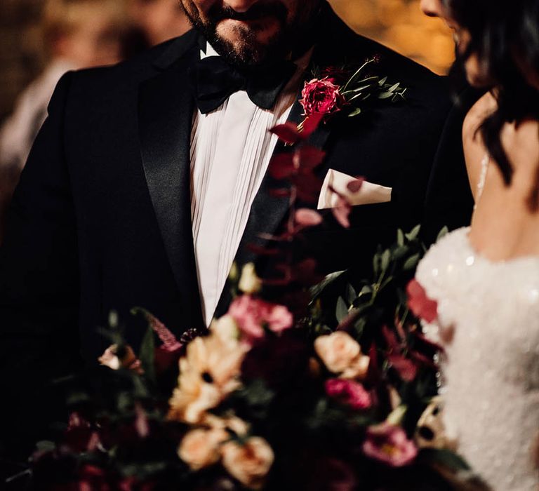 Smiling groom in black tie with a deep pink buttonhole smiling as the bride joins him at the altar 